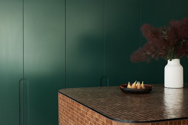 A close-up interior detail of a dark green built-in wall with fine vertical panel joints and discreet recessed pulls, paired with a substantial counter or island clad in small brown mosaic tiles. The counter has a dark edged surface and holds a white ceramic vase with dried burgundy foliage and a shallow bowl of decorative objects. The composition is moody, refined, and materially focused.