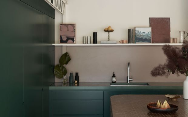 A moody, refined kitchen interior featuring deep green full-height cabinetry, matching lower cabinets, a dark stone benchtop, and a simple integrated sink with a tall brushed-metal tap. A pale wall-mounted open shelf displays books, ceramics, artwork and decorative objects, while a textured dark dining or kitchen table occupies the foreground. The styling combines muted natural tones, sculptural decor and dried foliage.