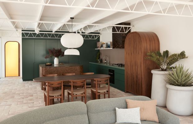 A warm, open-plan living and dining space with a dark green kitchen, curved timber cabinetry, a round timber dining table and chairs, and a pale curved sofa in the foreground. White exposed trusswork spans the ceiling, while patterned light flooring, sculptural pendant lights, indoor plants, and a softly glowing rounded doorway add distinctive character.