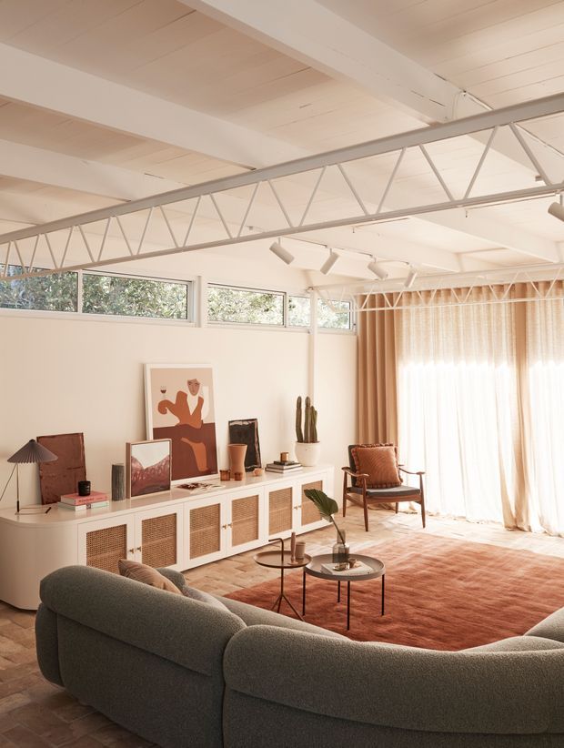 Bright, warm living room with a large curved upholstered sectional, mid-century-inspired lounge chair, low round coffee tables, textured area rug, and a long white built-in sideboard with woven-front cabinetry. The room features expansive windows with sheer curtains, white painted timber ceiling boards, exposed white-painted roof trusses, clerestory glazing, track lighting, and a relaxed art-filled interior.