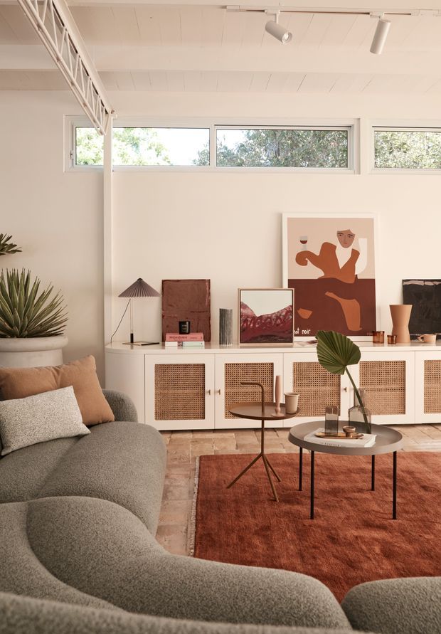 Warm, styled living room with a curved grey sectional sofa, woven cane-front low cabinetry, terracotta rug, round coffee tables, framed abstract artwork, indoor plants, and high clerestory windows beneath a white painted timber ceiling. The space has a refined mid-century-inspired look with soft earthy tones and abundant natural light.