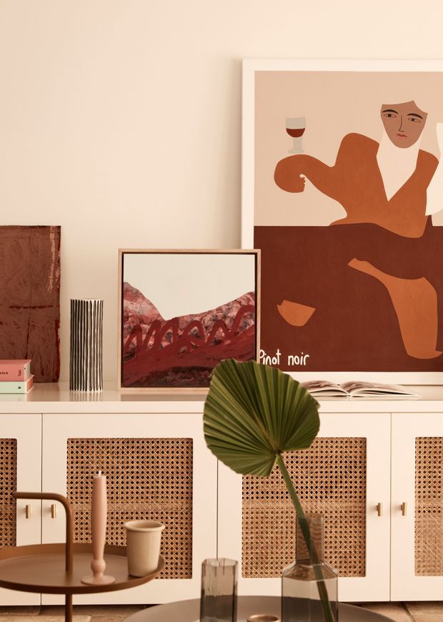 Warm, curated interior vignette featuring a cream built-in sideboard with woven cane-panel cabinet doors, layered framed artwork, books, and decorative objects. A round coffee table with a tray, candleholders, glassware, and a tall vase with a palm leaf occupies the foreground. The palette is earthy and muted, with terracotta, rust, cream, tan, and olive-green accents.