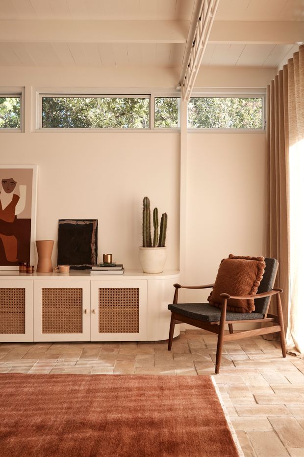 Warm, sunlit living room with a low white built-in sideboard featuring woven cane-front doors, a mid-century-style wooden lounge chair, terracotta cushion, cactus, artwork, and books. High horizontal windows, exposed white-painted ceiling boards and beams, woven curtains, terracotta-toned rug, and light brick or terracotta tiled flooring create a relaxed, earthy interior.