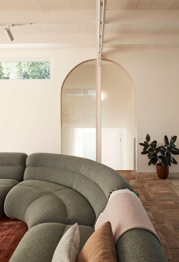 A softly lit contemporary living room with a large sculptural curved sectional sofa in muted green upholstery, layered neutral cushions, and a small potted rubber plant. The room features pale walls and ceiling, a warm parquet timber floor, an arched glazed doorway or opening, high-level horizontal glazing, and exposed ceiling-mounted track lighting. The overall palette is warm, muted, and organic with a refined contemporary feel.