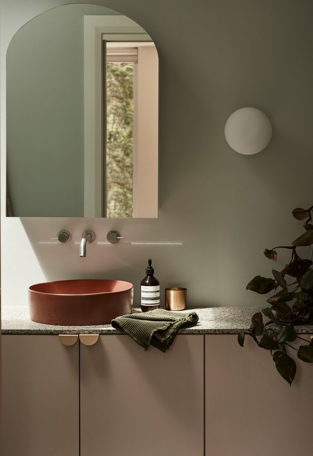 A warm, softly lit contemporary bathroom vanity featuring a terracotta vessel basin on a speckled stone benchtop, wall-mounted chrome tapware, pale muted green walls, a large rounded-corner mirror, and streamlined taupe cabinetry with small brass-toned pulls. A round wall light and trailing plant add sculptural and organic accents.