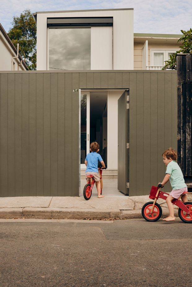 Street-level view of a contemporary residential frontage with a tall, box-like upper volume, large dark-framed window, and a muted vertical-panel fence enclosing a narrow entry gate. Two children with balance bikes are visible near the entrance. The facade combines pale rendered surfaces, horizontal weatherboard-style cladding, and dark timber fencing.
