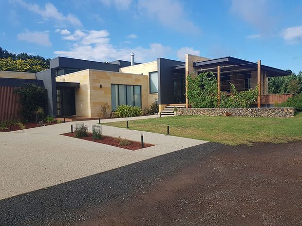 A contemporary single-storey residence viewed from the street, featuring low horizontal massing, flat rooflines, dark cladding, pale stone walls, large windows, and a sheltered timber-framed outdoor terrace with integrated greenery. The landscaped frontage includes a broad paved driveway, lawn, garden beds, and a low stone retaining wall.