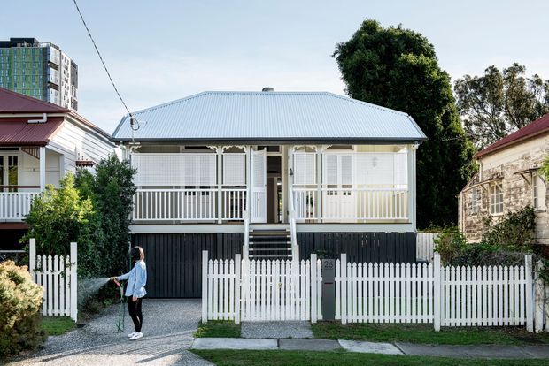 Front elevation of a compact Australian-style detached house with a raised verandah, pale corrugated metal hipped roof, white timber fretwork and balustrades, dark lower cladding, and a white picket fence. The home sits in an established suburban streetscape, with neighbouring buildings and mature trees visible around it.