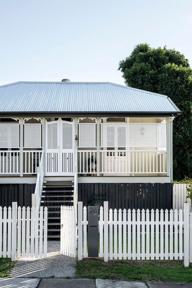 Front exterior view of a raised traditional timber cottage with a corrugated metal hipped roof, elevated enclosed verandah, decorative fretwork, white lattice screens, timber railings, dark lower cladding, and a white picket fence with front steps and gate. A leafy tree is visible behind the house.
