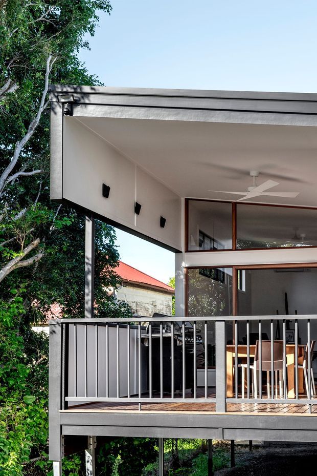 Exterior view of a raised contemporary house with a covered upper-level deck and open-plan dining area visible through extensive glazing. The structure features a broad flat roof or canopy, dark slender metal framing, pale rendered walls, timber-framed windows, and a grey metal balustrade. Dense mature vegetation surrounds the elevated deck, with a neighbouring red roof visible beyond.