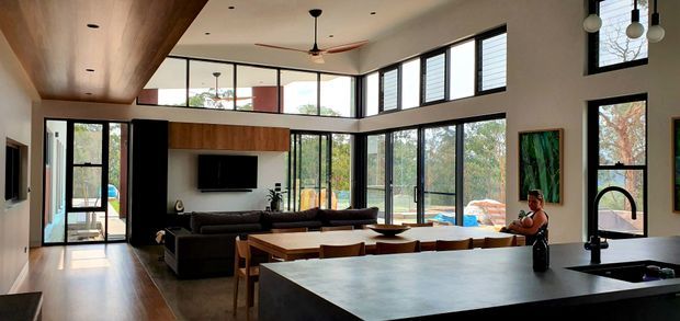 A spacious open-plan modern interior combining a kitchen, dining area and living room. The room features extensive black-framed floor-to-ceiling glazing, warm timber flooring and ceiling panels, a large dark kitchen island with sink, integrated living room television, timber dining table, and a high ceiling with clerestory windows. Lush trees are visible outside through the glazing.