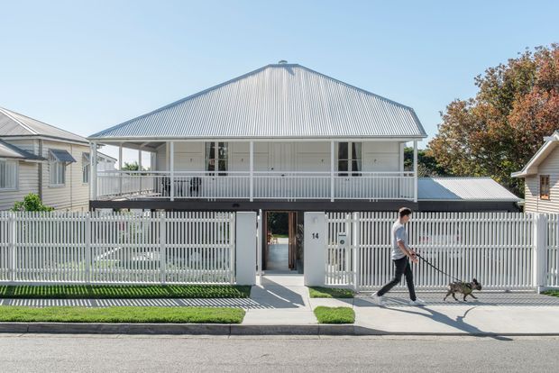 Front exterior view of a raised, single-storey residential house with a broad corrugated metal gable roof, covered upper veranda, white-painted weatherboard walls, and a tall white picket-style fence enclosing the front garden. A central pedestrian gate and front entry are visible, with neighbouring homes and a tree in the background.