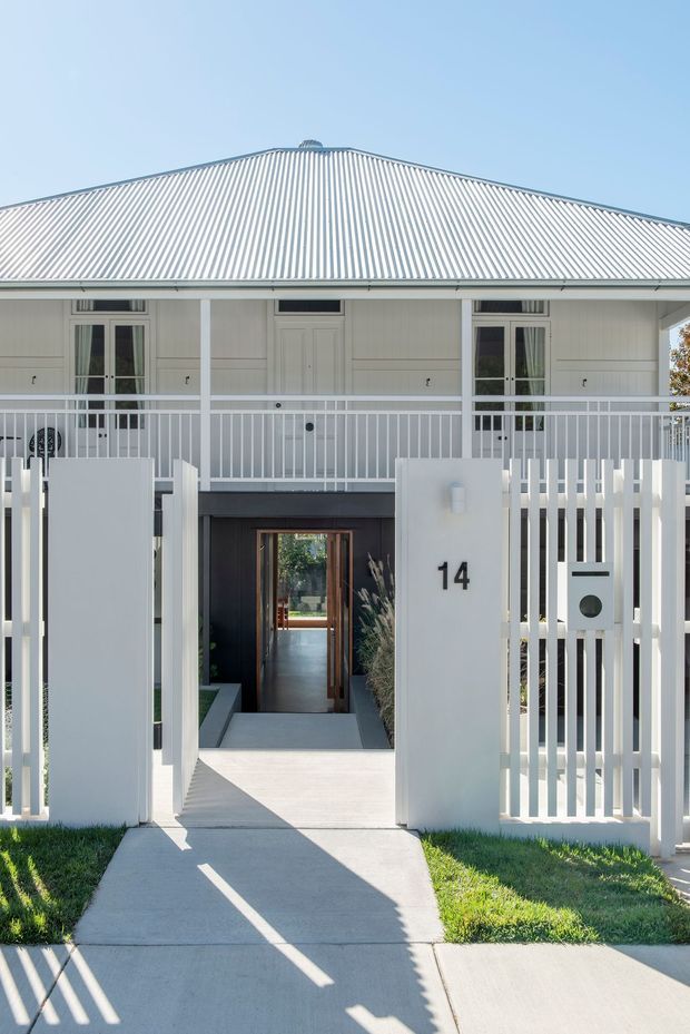 Front exterior view of a renovated light-coloured dwelling with a broad corrugated metal hipped roof, upper-level balcony/verandah with white balustrades, and a gated pedestrian entry. The lower entry is recessed in a dark charcoal wall, with a timber-framed glazed door, concrete path, white vertical-picket fencing, mailbox, and house number 14.