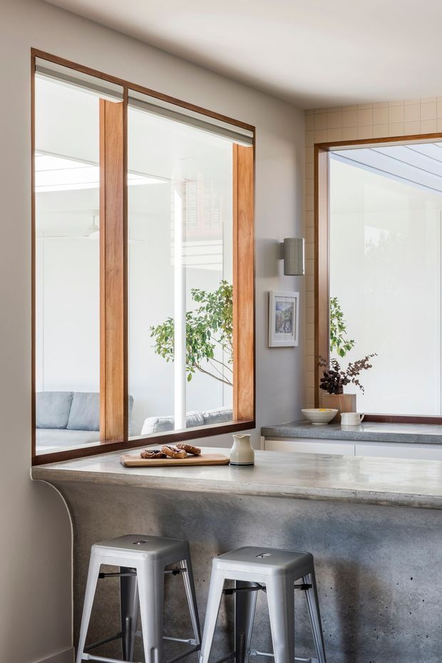 Bright contemporary kitchen interior with a substantial concrete island and integrated breakfast bar, paired with two metal stools. Large timber-framed glazed openings connect visually to a light-filled adjoining space, while a tiled wall section, pale cabinetry, stone-look counter surfaces, indoor plants and simple artwork create a clean, warm-modern atmosphere.
