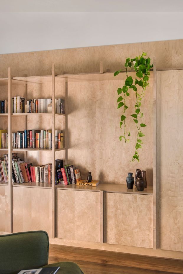 Interior view of a warm, compact living or study area with full-height light timber built-in shelving and cabinetry. The open shelves hold books, while a trailing indoor plant and dark ceramic vessels add decorative accents. A green upholstered chair is partially visible in the foreground, with timber flooring and a plain light ceiling completing the calm, natural scheme.
