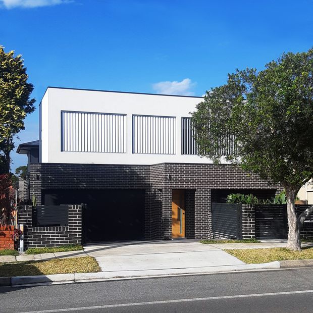 Street-facing contemporary two-storey residence with a crisp white upper volume, narrow vertical window screens, and a dark charcoal brick ground floor. The facade includes a large integrated garage, recessed timber entry door, dark horizontal-slat fencing, concrete driveway, and established street trees.