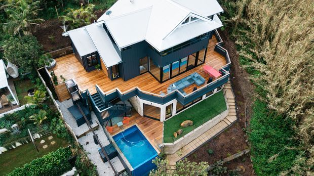 Aerial view of a multi-level contemporary home set into a lush, steeply landscaped site. The dark-clad house has a light pitched roof with triangular clerestory-like glazing, extensive timber decking, outdoor seating areas, terraces, stairs, and a bright blue swimming pool.