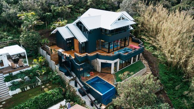 Aerial view of a multi-level contemporary house set into a lush, steeply vegetated site. The home has dark cladding, light metal pitched roofs with gabled sections, extensive glazing, multiple timber decks and balconies, and a rectangular swimming pool below. Landscaped gardens and a small detached outdoor structure are also visible.
