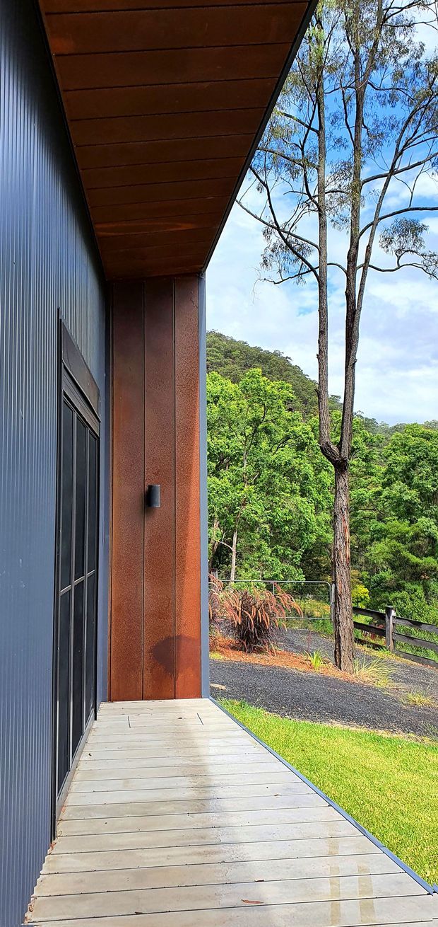A narrow covered exterior deck runs alongside a modern building entrance. The facade combines dark vertical corrugated cladding with rust-toned metal wall panels, a timber-lined soffit, and a dark glazed entry door. The deck is finished in pale timber, opening toward a lush wooded landscape with native-style planting and a gravel path.