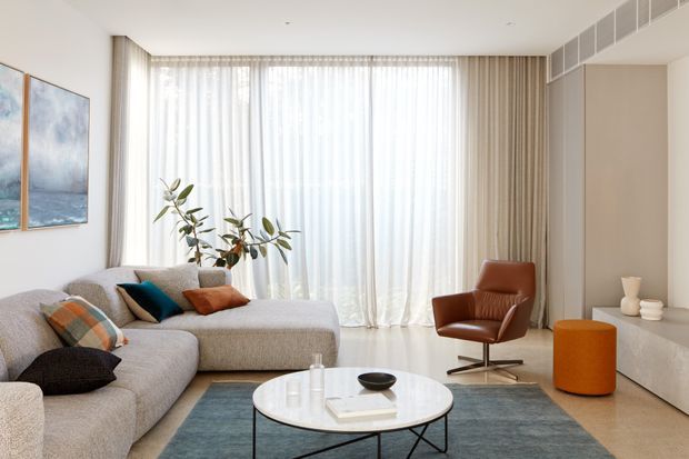 Bright contemporary living room with a large L-shaped upholstered sectional, leather lounge chair, round white coffee table, and orange cylindrical ottoman. Full-height sheer curtains span the main window, with neutral walls, vertical drapery, a muted blue-grey rug, framed artwork, and restrained styling creating an airy, refined interior.