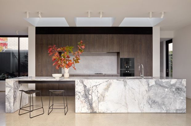 A refined contemporary kitchen centred on a large waterfall island clad in heavily veined pale natural stone. Dark timber-look cabinetry forms a full-height feature wall with an integrated cooktop and appliances, while a pale stone benchtop continues across the island and rear work surface. Minimal white geometric pendant lights, slim black-framed glazing, and two simple black bar stools complete the clean, high-contrast composition.