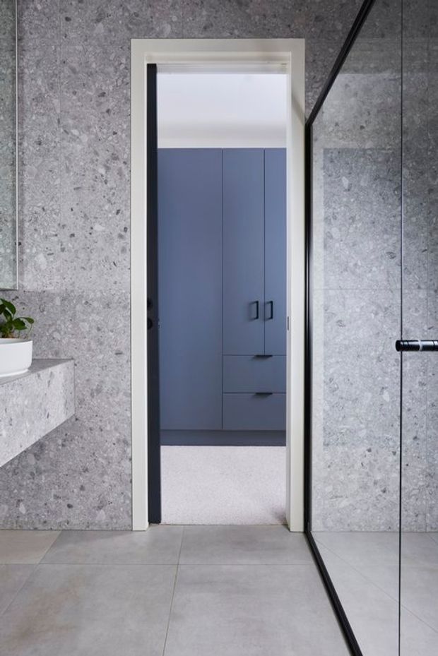 A minimalist bathroom interior with large grey stone-look wall and floor tiles, a frameless glass shower screen with black hardware, and a white-trimmed doorway opening to built-in blue-grey cabinetry in an adjacent space. A floating stone ledge with a small plant is visible at left.