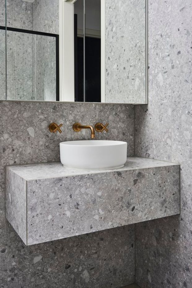 Close-up of a refined bathroom vanity featuring a white vessel basin, wall-mounted brass-gold tapware, mirrored cabinet, and a floating vanity clad in grey terrazzo or stone with matching wall tiles.