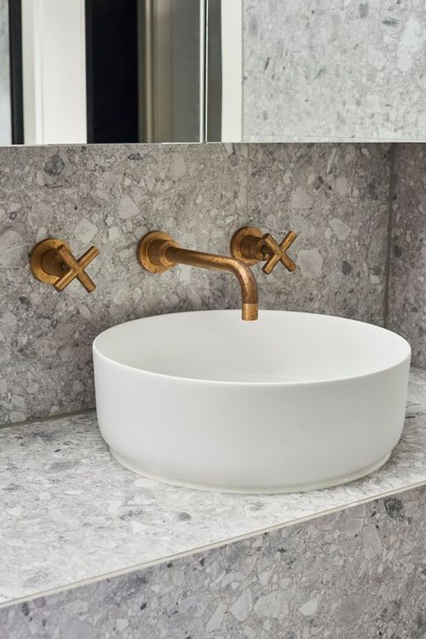 Close-up of a refined bathroom vanity featuring a white round vessel basin on a grey veined stone benchtop, with a matching grey stone wall surface and exposed cross-handle wall-mounted tapware in a warm bronze or brass finish. A mirror is visible above the basin.
