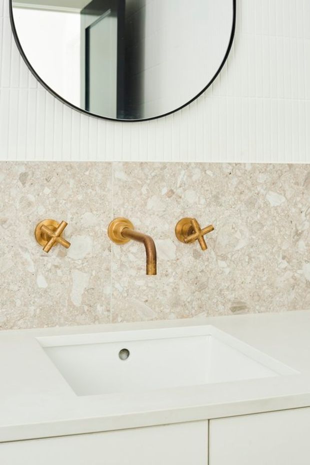 Close-up of a refined bathroom vanity featuring a white integrated rectangular basin, wall-mounted brushed gold cross-handle tapware, a pale natural stone splashback, white vertically textured wall tiles, and a large round black-framed mirror.