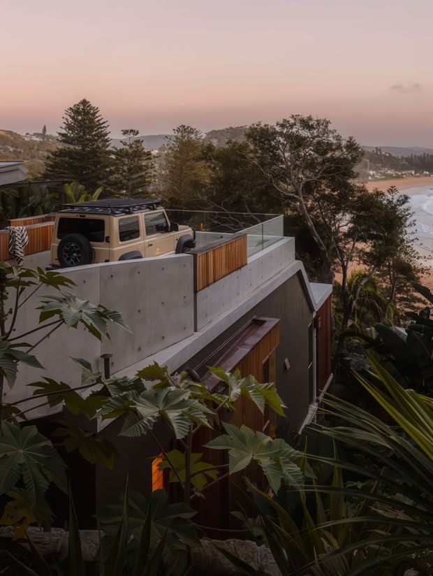 A dusk exterior view of a multi-level contemporary coastal house set into a steep, heavily landscaped site. The building features exposed concrete walls, dark cladding, warm timber screening, rooftop terraces with glass balustrades, and a parked vehicle on an upper deck. Beyond the trees is a sandy beach, ocean, and rugged coastal headland under a pink sky.