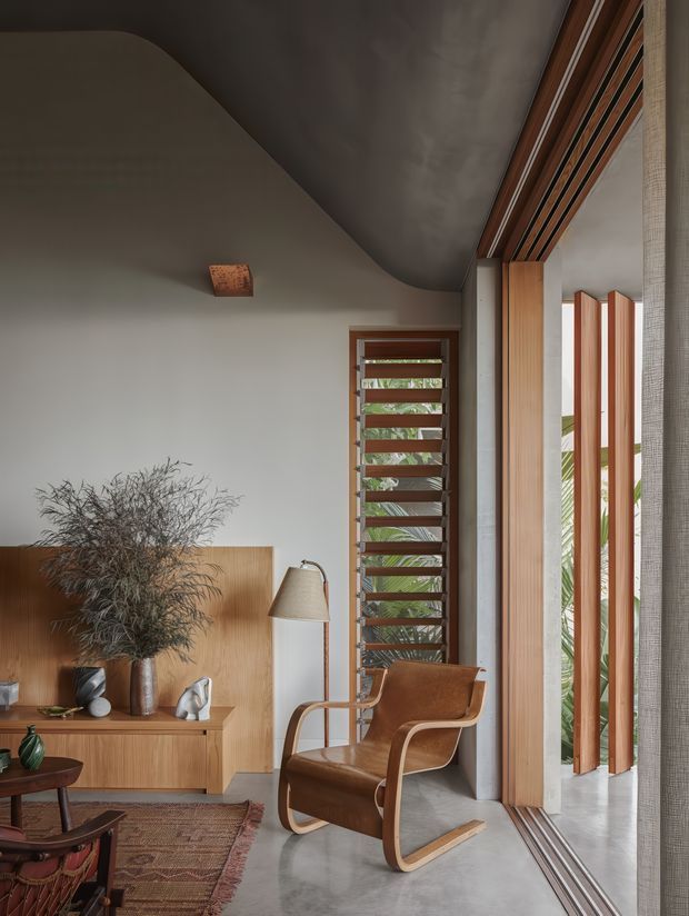 A warmly styled living room with a sculptural curved ceiling, polished concrete floor, timber cabinetry, and extensive timber-framed glazing with vertical external sun-shading fins. A brown leather-and-bentwood rocking lounge chair sits beside a floor lamp, with a low timber media console, patterned rug, and natural branch arrangement adding texture. The space has strong indoor-outdoor connections and a refined mid-century-influenced contemporary character.