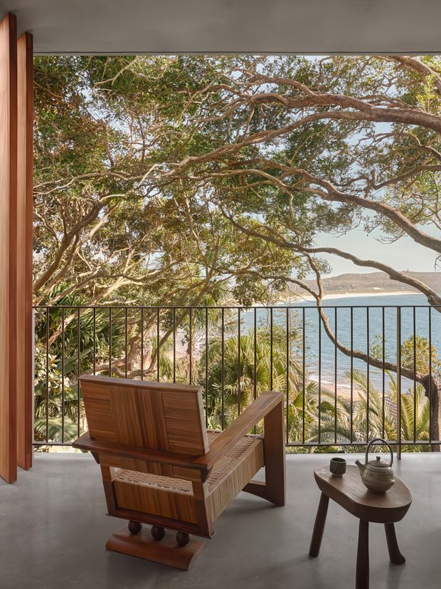 A serene indoor-outdoor sitting area opens onto a balcony or terrace overlooking a tropical, tree-lined bay. The space features a sculptural timber lounge chair, a small dark wood side table with a ceramic teapot and cup, polished concrete flooring, warm timber door or wall framing, and slender black metal balcony railings. Dense branches frame the water view, with a sandy shoreline and hills visible in the distance.