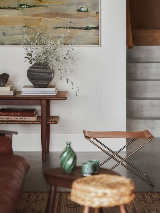 A warmly styled living room vignette with a dark timber console table, decorative books, a large ceramic vase of dried foliage, framed abstract artwork, and a leather lounge chair. A low wooden coffee table with green glass objects sits over a patterned rug, while a timber-and-metal folding lounge chair occupies the right side. The room combines natural materials, earthy tones, and mid-century-inspired furnishings with subtle wall panelling and a partially visible textured architectural element.