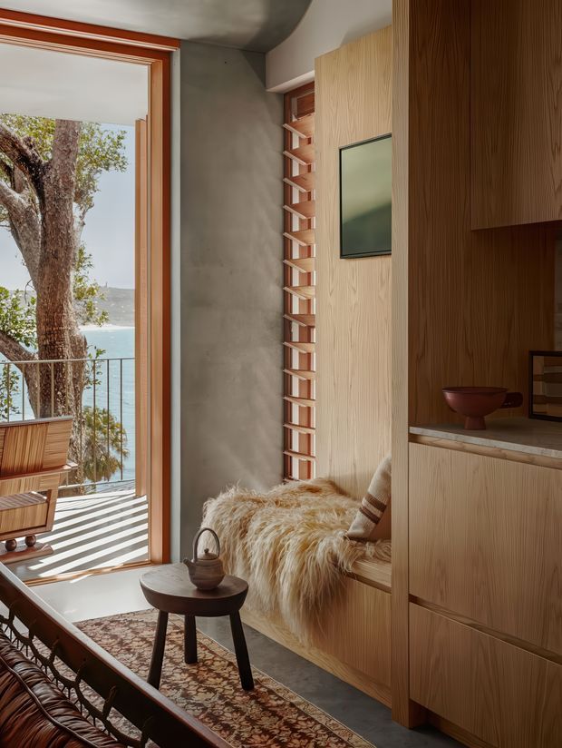 A warm, compact living room with a built-in timber-lined window seat, vertical wood cabinetry, and a small dark wood side table. Full-height glazing opens to a narrow balcony with views of water and trees. The room features a textured concrete wall, timber window frames, patterned runner rug, leather seating, and layered natural textiles, creating a refined mid-century-inspired retreat.