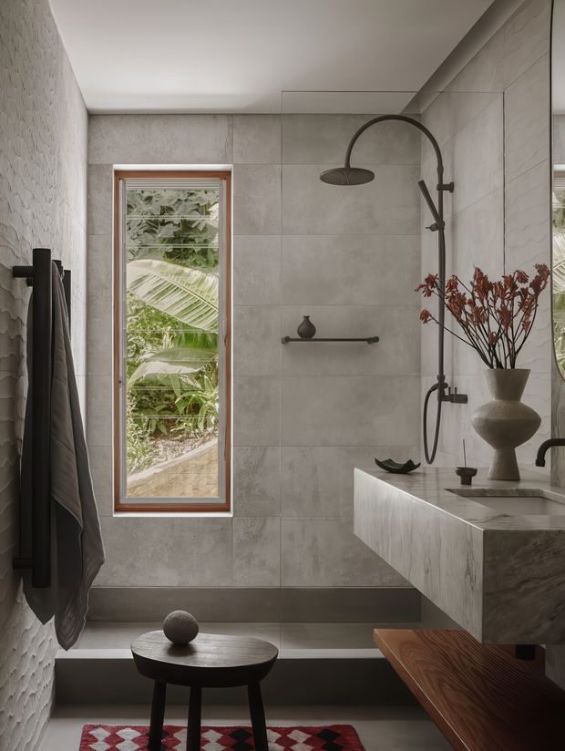 A refined contemporary bathroom featuring a walk-in shower with a dark metal rainfall shower system, large grey stone-look wall tiles, a floating stone vanity, and a narrow timber-framed window overlooking lush greenery. Sculptural accessories, a dark timber stool, and a patterned rug add warmth and character.