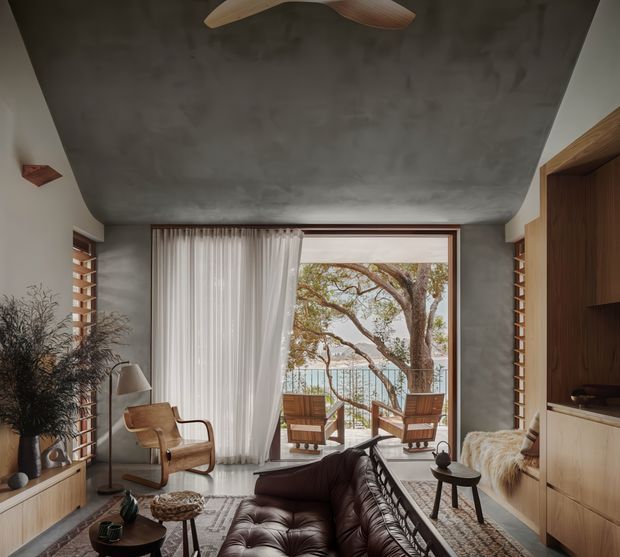 Warm, refined living room with a low, sculptural concrete-look ceiling, timber built-in cabinetry, and large glazed doors opening to a balcony with a tree and water view beyond. The room contains a dark tufted leather sofa, mid-century-style timber lounge chairs, sheer curtains, a patterned rug, and layered decorative lighting. The composition emphasizes the indoor-outdoor connection and natural timber detailing.