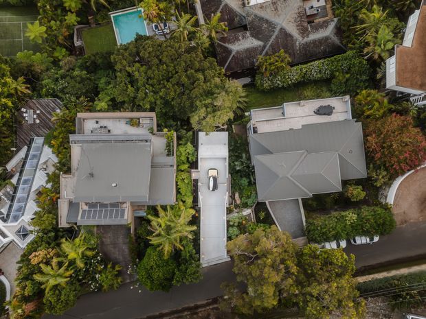 Aerial overhead view of a leafy residential neighbourhood showing several detached homes, varied roof forms, driveways, dense tropical landscaping, and a turquoise swimming pool. The image is dominated by the building exteriors and surrounding suburban site rather than any single interior space.