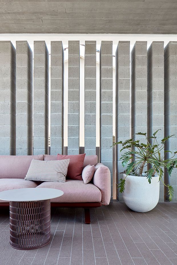 Interior living room vignette featuring a pale pink upholstered sofa, patterned cushions, a sculptural round coffee table, and a large potted plant. The space is defined by repeated vertical grey concrete block piers with narrow openings, a dark tiled floor, and a timber-lined ceiling. The composition has a refined contemporary character with strong textural and geometric elements.