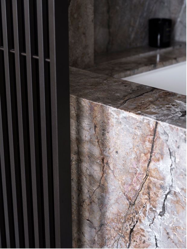 Close-up architectural detail of a luxurious bathroom, showing a thick natural stone bath surround or benchtop with dramatic veining, a dark vertical metal screen or radiator at left, and a white bathtub partially visible in the background.