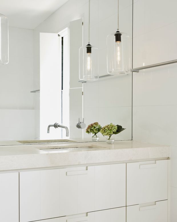 Bright, minimalist bathroom vanity with a wide white built-in cabinet, pale stone benchtop, integrated basin and chrome wall-mounted tapware. Large mirrored wall panels reflect the space, while clear cylindrical pendant lights hang above the vanity. A small arrangement of green foliage adds a natural accent.