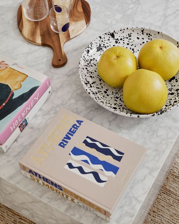 A styled tabletop vignette on a pale grey-white marble surface, featuring a black-speckled white bowl filled with yellow grapefruits, a wooden serving board with glasses, and stacked art and design books. A woven rug is partially visible at the edge, creating a warm contemporary still-life composition.