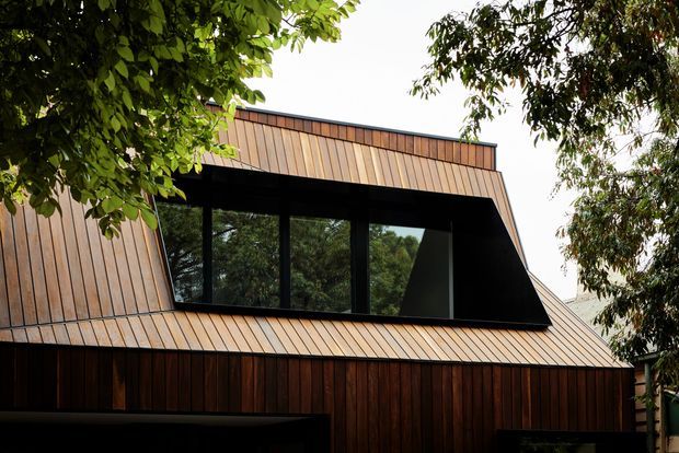 Exterior architectural photograph of a contemporary, angular house partially framed by mature trees. The facade and steeply folded roof are clad in warm vertical timber boards, with a large dark-framed horizontal window band recessed beneath an overhanging roof form. The composition emphasizes the unusual sloped roofline, strong geometric massing, and contrast between dark glazing and rich timber cladding.