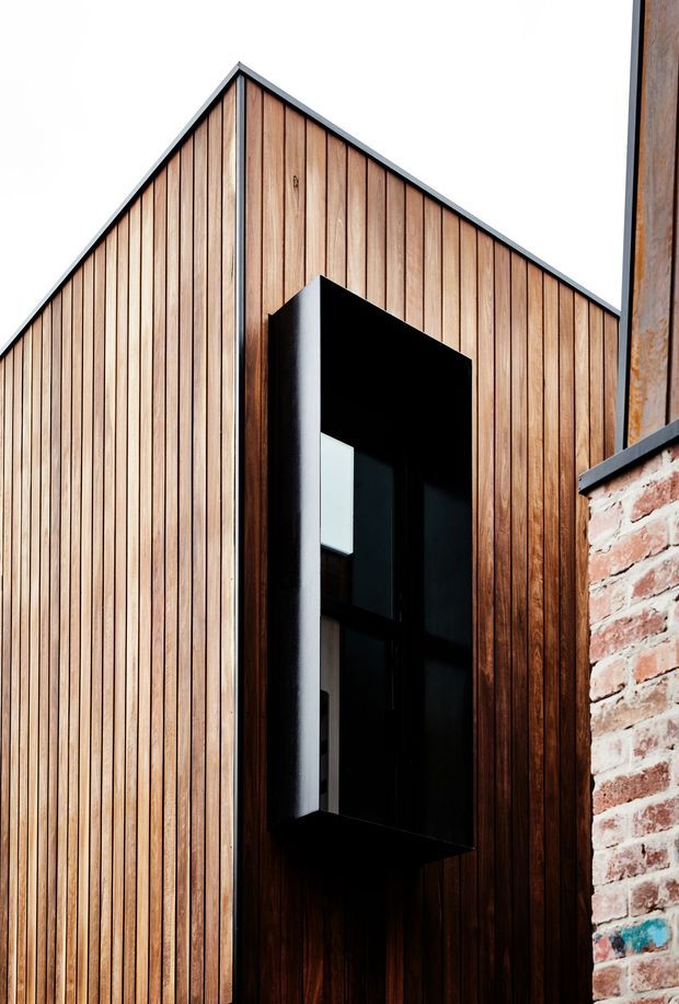 Close-up exterior view of a contemporary building facade with a steep gabled roofline, vertical warm timber cladding, dark metal edging, and a tall black-framed recessed window or opening. A weathered exposed brick wall is visible at the right edge, creating a contrast between new timber construction and existing masonry.
