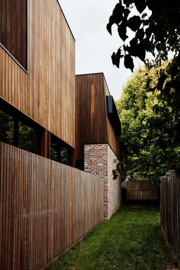 A narrow landscaped side passage beside a contemporary multi-level building. The facade and tall boundary fencing are clad in vertically arranged timber boards, with dark-framed glazing beneath projecting upper volumes. A contrasting exposed red brick wall forms part of the lower structure, while mature greenery encloses the passage and a timber gate is visible at the far end.