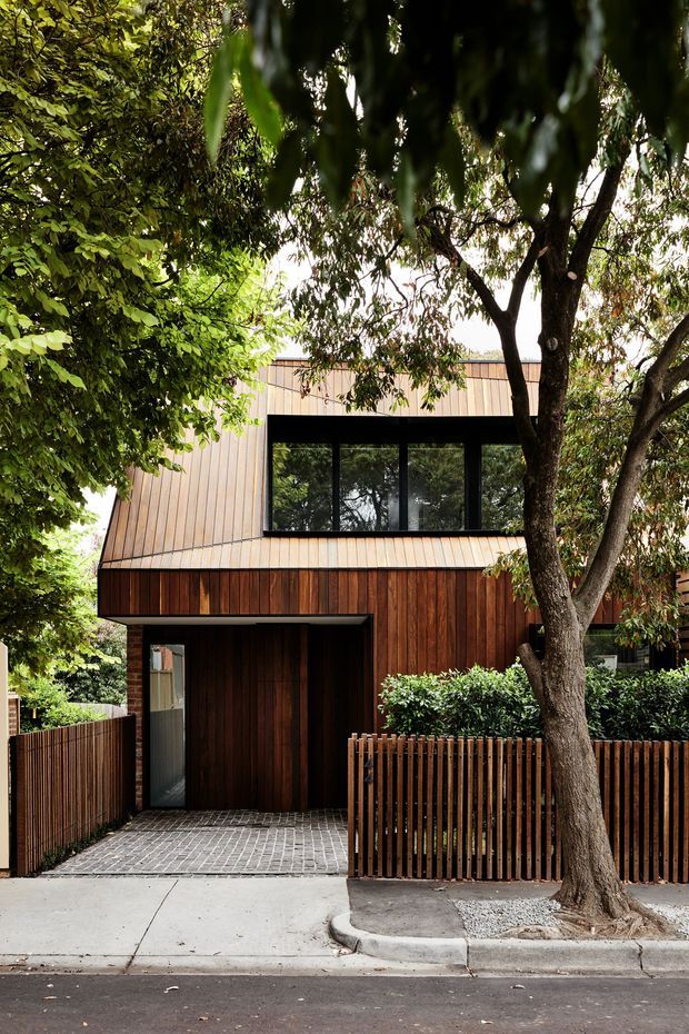 Street-facing view of a compact contemporary two-storey residence, largely clad in warm vertical timber boards with dark-framed upper-level glazing. A recessed entry and paved driveway sit behind a timber slat fence, while mature trees and dense planting frame the facade.