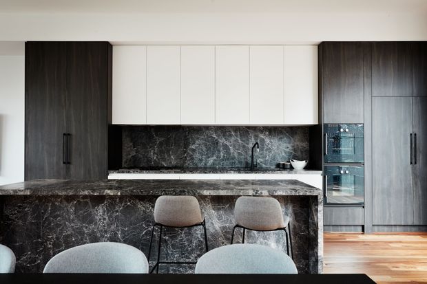 Contemporary kitchen with a large dark stone island and matching splashback, dark timber-toned cabinetry, white overhead cupboards, integrated stainless-steel appliances, and a black tap. Two bar stools sit at the island, with additional seating in the foreground. Warm timber flooring contrasts with the monochromatic cabinetry and stone surfaces.