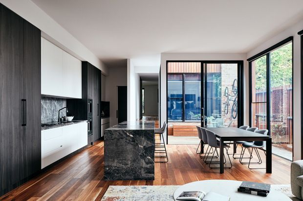Open-plan contemporary kitchen and dining area with dark timber and white cabinetry, a dramatic dark stone island, warm timber flooring, and extensive black-framed glazing. The space opens to a bright outdoor area and includes a compact dining table with modern chairs.