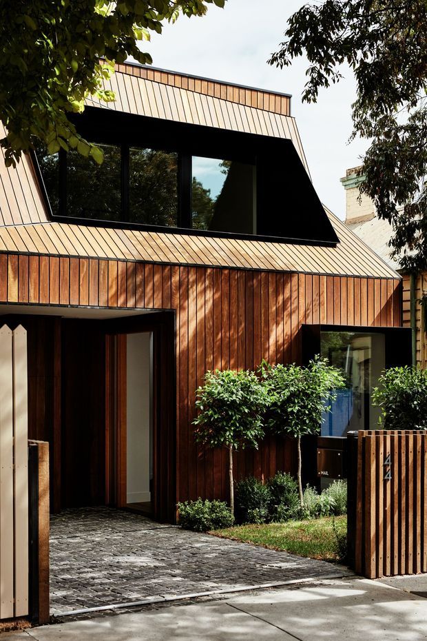 Street-facing view of a compact contemporary urban house with a strongly geometric form. The facade and steep roof planes are clad in vertically aligned warm timber, with dark-framed glazing, a recessed upper-level window, a covered entry, paved driveway, and timber slatted front fence. Mature planting partially frames the building, while a neighbouring masonry structure is visible at the side.