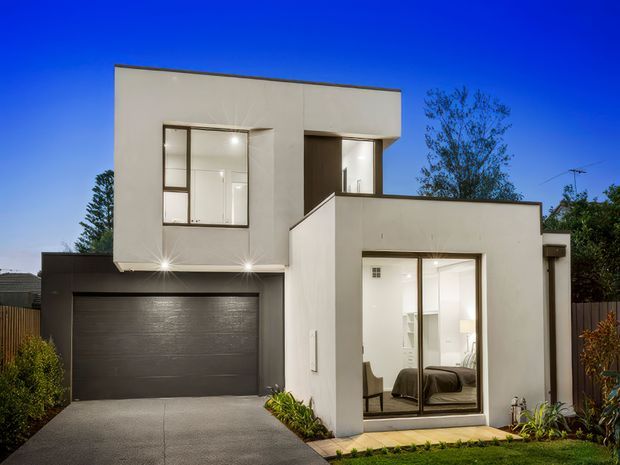 A contemporary multi-level residential home viewed from the street at dusk. The minimalist white rendered facade features clean geometric volumes, large windows, a dark integrated double garage, and a ground-floor bedroom or living space opening toward a small landscaped front garden. Warm interior lighting contrasts with the deep blue sky.