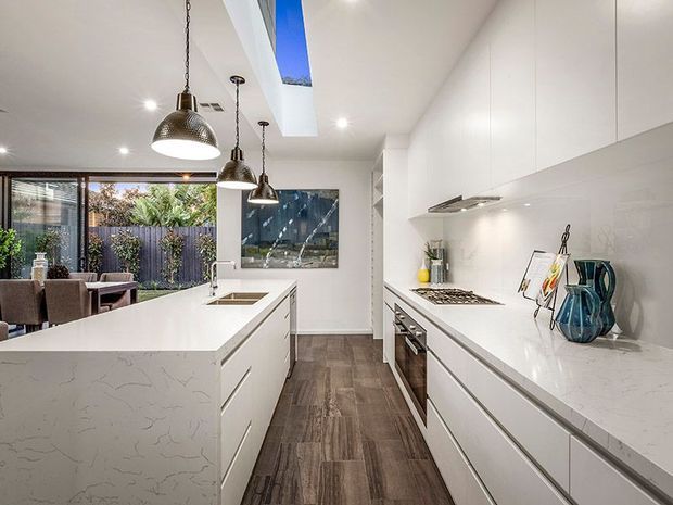 Bright contemporary galley-style kitchen with a large white waterfall island, extensive white cabinetry, integrated appliances, pendant lighting, and a skylight. Dark wood-look flooring contrasts with the light cabinetry, while full-height glazing connects the kitchen to an outdoor dining area and garden.
